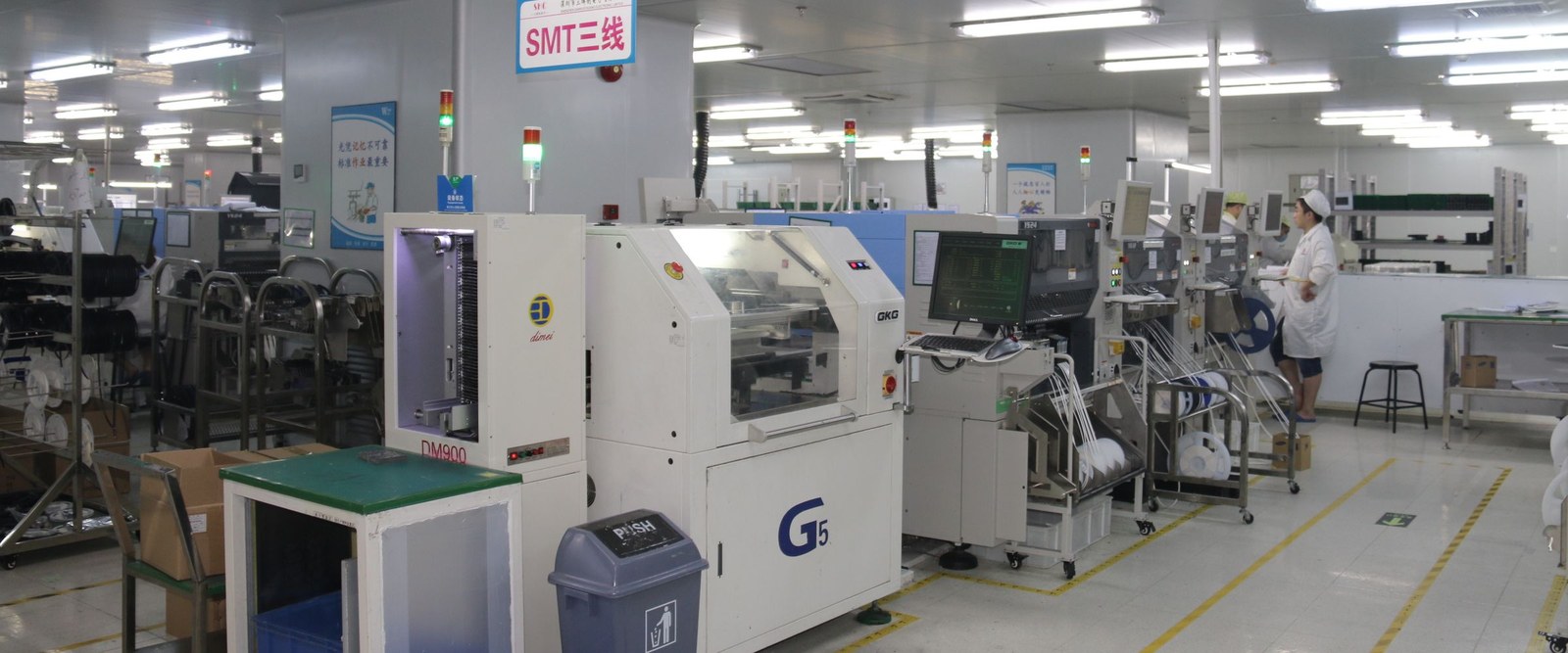 A white SMT line: a screen printer and a placement machine along a conveyor, two carts loaded with component reels, an operator in a white coat, and yellow floor markings